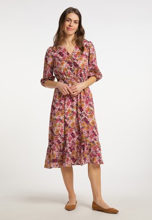Vrouw staand in een bloemenprint midi-jurk in roze, oranje en paars met driekwart mouwen en bruine platte schoenen tegen een effen witte achtergrond.