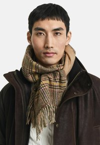 Junger Mann mit kurzen schwarzen Haaren, der einen braun karierten Schal, einen weißen Strickpullover und eine dunkelbraune Lederjacke vor einem weißen Hintergrund trägt.