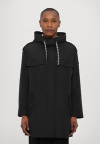 Parka noire avec capuche, dotée d'une fermeture éclair à l'avant, de deux poches poitrine et d'un cordon de serrage. Texture lisse et coupe légèrement oversize.