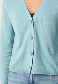Gilet bleu clair avec un col en V, cinq boutons gris et un ourlet côtelé, fabriqué à partir d'un tissu doux et texturé. Idéal pour le superposition.