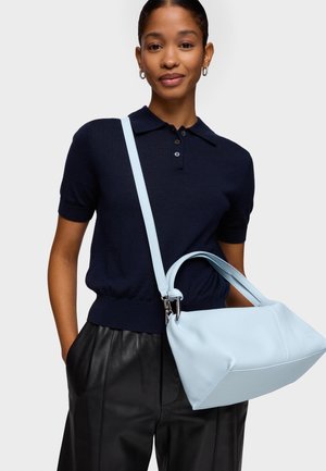 Mujer que lleva un polo de manga corta azul marino y pantalones negros, sosteniendo un bolso de cuero azul claro con una correa para el hombro cruzada sobre su cuerpo.