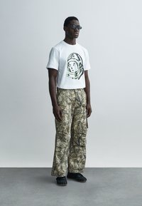 Λευκό κοντομάνικο t-shirt με γραφικό σχέδιο, σε συνδυασμό με μπεζ καμουφλάζ παντελόνι cargo και μαύρα υποδήματα. Φοράει γυαλιά ηλίου και στέκεται όρθιος.