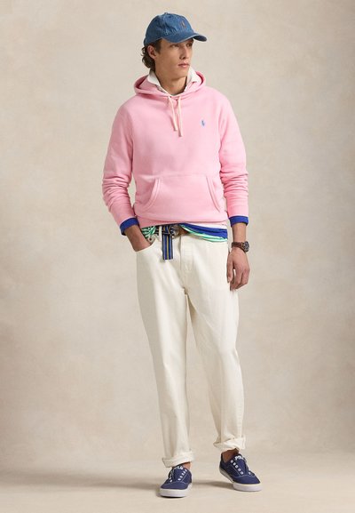 Jaunas vyras su rožiniu gaubtu, mėlyna kepure, baltais kelnėmis, dryžuotu diržu ir tamsiai mėlynomis sportbačiais stovi kišenėse rankas laikydamas ir žiūri į šoną.