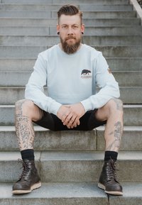 Sudadera azul claro con logo de oso, pantalones cortos negros, botas de cuero marrón oscuro con cordones. Tatuajes visibles en las piernas, sentado en escaleras de piedra.