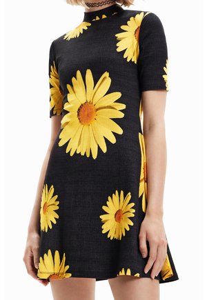 Robe noire à manches courtes avec de grands motifs de marguerites jaunes, texture côtelée, et col montant ajusté portée par une personne debout.