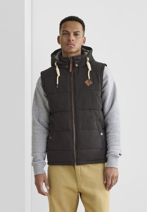 Padded black vest with hood, zip closure, and beige drawstrings. Worn over a light gray long-sleeve shirt, paired with beige pants.