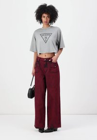 Grå kort t-skjorte med "GUESS"-logo, kombinert med vide maroon denimbukser med frontlommer og subtil tekstur. Svart håndveske inkludert.