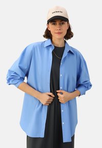 Chemise boutonnée bleu clair avec une coupe décontractée, des manches longues et un ourlet arrondi. Portée par-dessus un haut sombre. Une casquette beige avec des lettres noires complète le look.