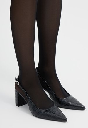 Gambe con collant neri trasparenti che indossano scarpe nere con tacco a blocco, punta affilata e texture effetto coccodrillo, con cinturino posteriore, su sfondo bianco.