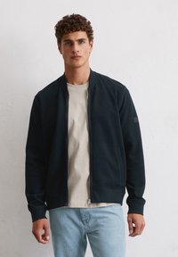 Navy zip-up bomber jacket with ribbed cuffs and collar, worn over a light beige shirt and paired with light blue jeans. Smooth texture.