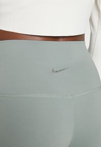 Svetle zelené sportovní legíny vyrobené z elastického materiálu, s malým, třpytivým stříbrným logem Nike v pravém horním rohu.