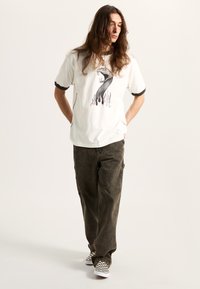 Camiseta blanca con ribete negro, que presenta un gráfico de una serpiente rodeada de llamas coloridas. Combinada con pantalones cargo olivo y zapatillas de cuadros.