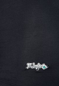 Svart figursydd tygplagg med en liten silverlogotagg med texten "Xhuja" och en turkos accent, som betonar en slank design och en mjuk textur.