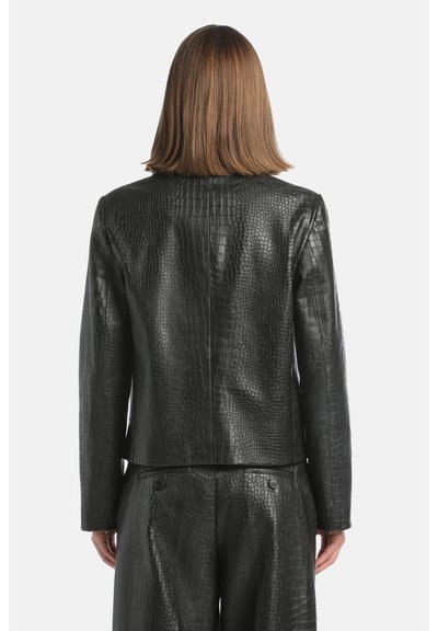 Persoon met schouderlang lichtbruin haar, gekleed in een zwart faux krokodillenleren jack en bijpassende broek, gezien van achteren.