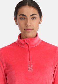 Rosa Fleece-Pullover mit hohem Kragen, halbem Reißverschluss und einem dekorativen Spinnenlogo aus Strasssteinen, das in der Mitte der Brust platziert ist.
