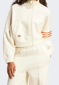 Krämfärgad zip-up sweatshirt med hög krage, med röda tre-randiga detaljer på ärmen och broderad logotyp på fickan.