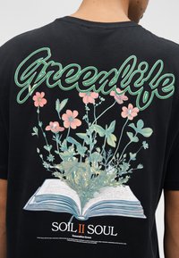 Spate cămașă neagră cu textul „Greenlife” deasupra unei cărți deschise din care cresc plante verzi și flori roz, iar dedesubt „Soil II Soul Generation Green”.