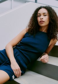 Woman with curly hair wearing a sleeveless navy dress, reclining on concrete stairs indoors.