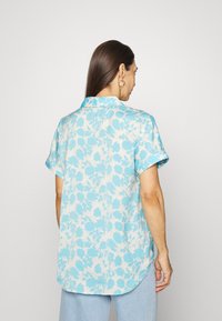 Camisa de mangas curtas em azul claro com um padrão floral, feita de tecido macio. Apresenta uma bainha curva e um colarinho pontiagudo.