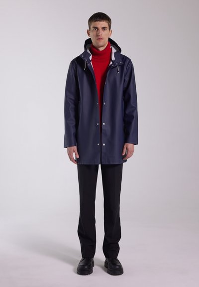 Navy waterproof jacket with hood, featuring snaps, side pockets, and a smooth texture, worn over a red turtleneck and black pants.