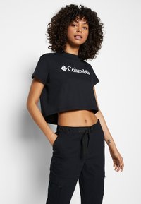 T-shirt cropped nera con logo bianco "Columbia". Realizzata in cotone, con maniche corte e taglio rilassato. Indossata con pantaloni cargo scuri.