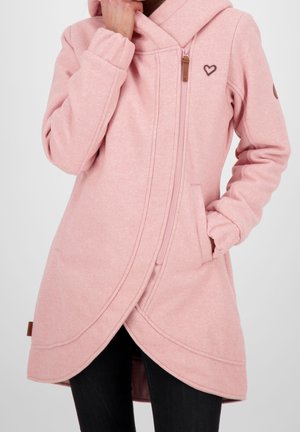 Femme portant une longue veste à capuche rose avec une fermeture éclair asymétrique à l'avant et un petit emblème en forme de cœur sur la poitrine.