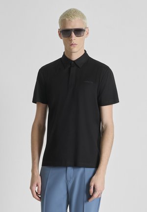 Joven con cabello rubio corto que lleva gafas de sol oscuras, una camiseta polo negra y pantalones azul claro frente a un fondo gris liso.