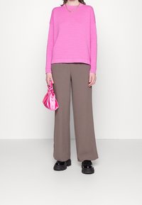 Kvinna iklädd en rosa ribbad tröja, bruna vida byxor, svarta klumpiga skor, guldchainhalsband och håller i en rosa satin scrunchie-väska.