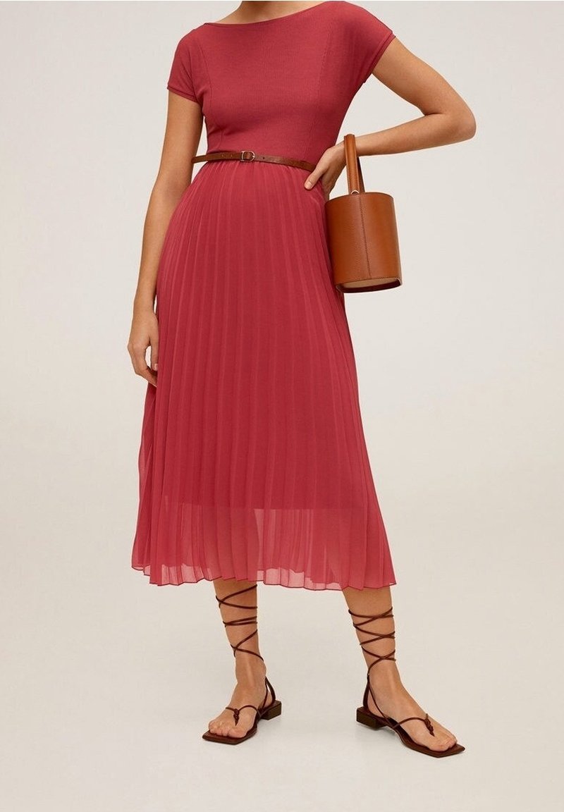 Femme en robe midi plissée rouge avec ceinture marron, tenant un sac à main cylindrique marron, portant des sandales plates marron à lacets.
