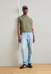 Olive green short-sleeve t-shirt, light blue jeans, black sneakers with white stripes, gray cap, standing against a light background.