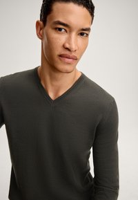 Pullover lungo verde scuro con maniche a lungo e scollatura a V, con una texture liscia e un design aderente, caratterizzato da dettagli di cucitura lungo il collo.