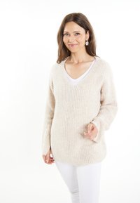 Pulover bej cu nervuri, cu guler în V și mâneci lungi, asortat cu o cămașă albă și pantaloni albi, având o croială lejeră și o textură moale.