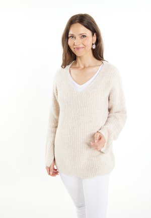 Beige geribbelde trui met een V-hals en lange mouwen, gecombineerd met een wit shirt en witte broek, met een ontspannen pasvorm en zachte textuur.