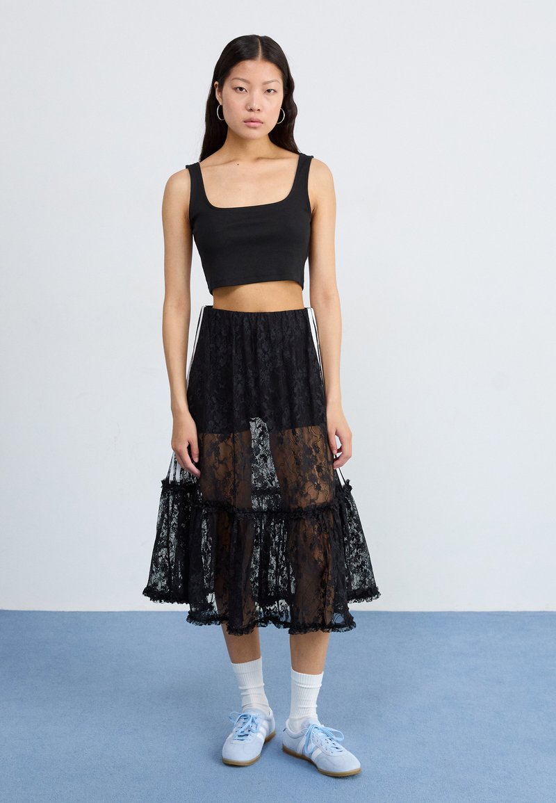 Junge Frau mit langen Haaren trägt ein schwarzes Crop-Top und einen durchsichtigen schwarzen Spitz-Rock, steht auf einem blauen Boden vor einem hellgrauen Hintergrund.