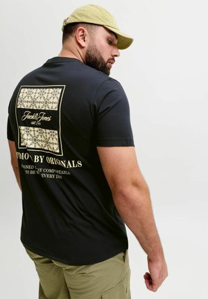 Skæggede mand iført en sort T-shirt med grafik og beige kasket, der kigger over sin skulder, med khakibukser mod en ensfarvet lys baggrund.