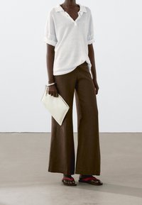 Leichtes weißes Polo mit aufgerollten Ärmeln, kombiniert mit lockeren braunen weiten Hosen. Hält eine cremefarbene Clutch, trägt schwarze und rote Flip-Flops.