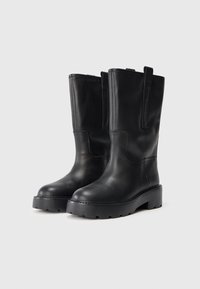 Botas altas de cuero negro hasta la rodilla con punta redonda, suela de goma gruesa y tiradores en los lados para facilitar su uso. Textura suave y costuras cosidas.