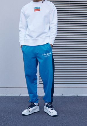 Sudadera blanca con un logo "TOMMY Jeans" en rojo y verde, combinada con pantalones de chándal azules que llevan "New York" en escritura blanca. Zapatillas blancas con detalles en negro.