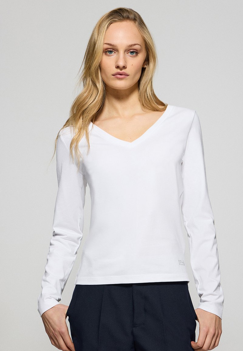 Maglietta bianca a maniche lunghe con scollo a V realizzata in tessuto morbido, caratterizzata da un logo del marchio in basso a sinistra. Design classico e aderente, semplice e pulito.