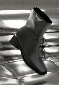 Fekete bőr bokacsizma négyzet alakú orral, alacsony sarokkal és fémes kiegészítővel a bokán. Simább textúra, minimalista dizájn.