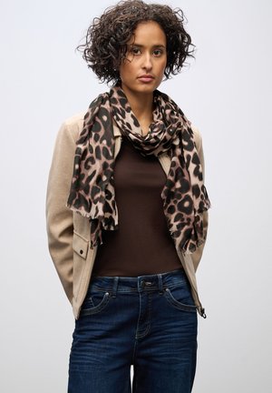 Giacca in suede beige sopra una maglia marrone, con una sciarpa a stampa leopardata. Jeans blu scuro con tasche, indossati all'altezza della vita. Capelli corti e ricci.