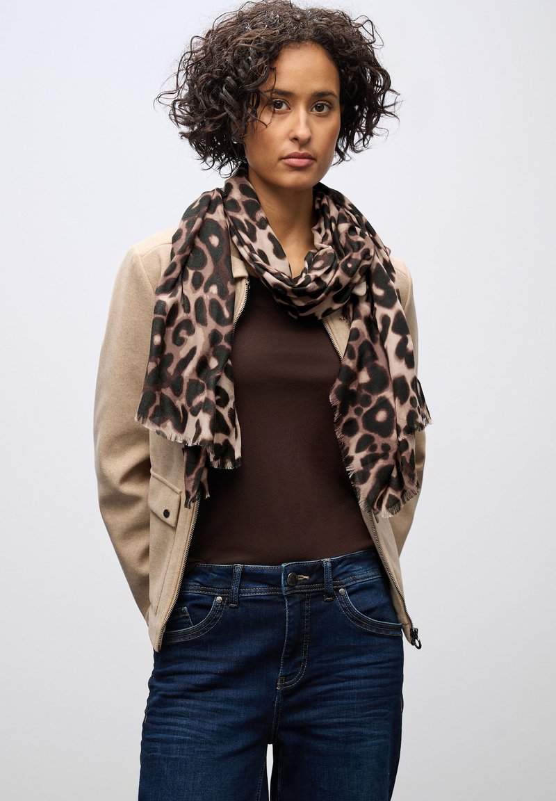 Beige mockajacka över en brun topp, med en scarf i leopardmönster. Mörkblå jeans med fickor, bärs vid midjehöjd. Kort lockigt hår.