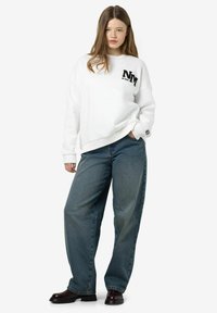 Witte sweatshirt met zwart geborduurd "NM" logo op de borst, gecombineerd met losse blauwe denim jeans en zwarte platformlaarzen.