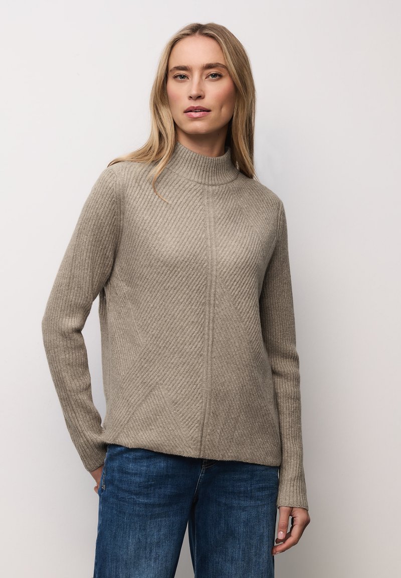 Femme portant un pull à col roulé côtelé beige et un jean bleu, debout devant un fond clair uni.