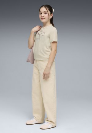 Niña joven con cabello oscuro largo, camiseta beige de Puma, pantalones anchos beige, zapatillas blancas, sosteniendo un bolso texturizado rosa sobre su hombro.