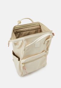 Sac à dos beige en tissu doux avec une fermeture zippée sur le dessus, deux poches latérales et des accents en métal doré. Intérieur doublé en tissu clair.