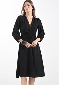 Abito nero al ginocchio con scollo a V, vita arricciata e maniche lunghe a sbuffo. Presenta un tessuto texturizzato e dettagli con cordoncino per la regolazione.