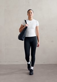 Witte sportt-shirt en zwarteleggings, gecombineerd met een zwarte schoudertas en zwarte sneakers, tegen een gladde, neutrale achtergrond.