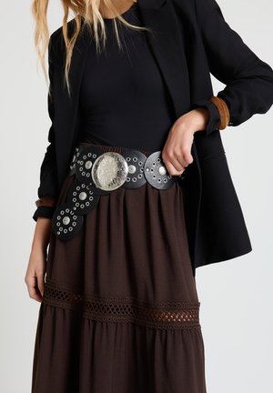 Frau trägt schwarzen Blazer und Oberteil mit braunem Rock, der Spitzenverzierung und einen breiten schwarzen Gürtel mit großen silbernen runden Verzierungen aufweist.
