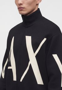 Homme portant une veste noire zippée avec de grandes lettres blanches "A", "X" sur le devant et les manches, col montant, raccourcie au niveau du cou et des épaules.